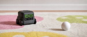A small charcoal grey Boxer robot toy on a patterned kids rug, its black face panel lit with a grid of bright green LED dots forming two blocky eyes, its white ball resting a short distance ahead of it.