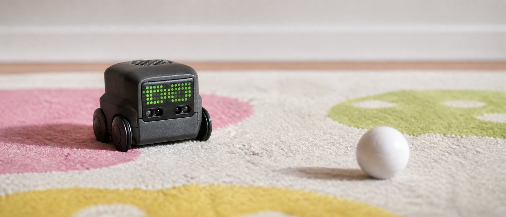A small charcoal grey Boxer robot toy on a patterned kids rug, its black face panel lit with a grid of bright green LED dots forming two blocky eyes, its white ball resting a short distance ahead of it.