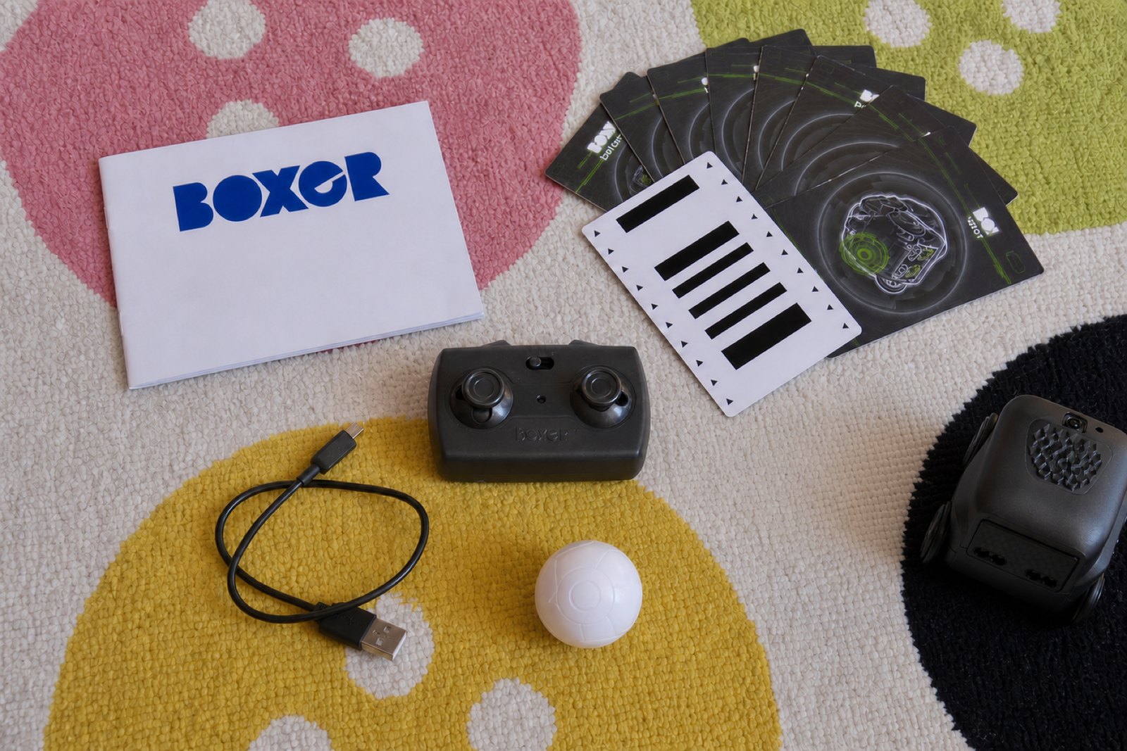 Overhead flat lay on a patterned rug of everything in the Boxer box: the robot, a charcoal remote with two thumbsticks, a white ball, a black Micro USB cable, a fanned stack of black activity cards, one card flipped to its striped white side, and a white instruction booklet.