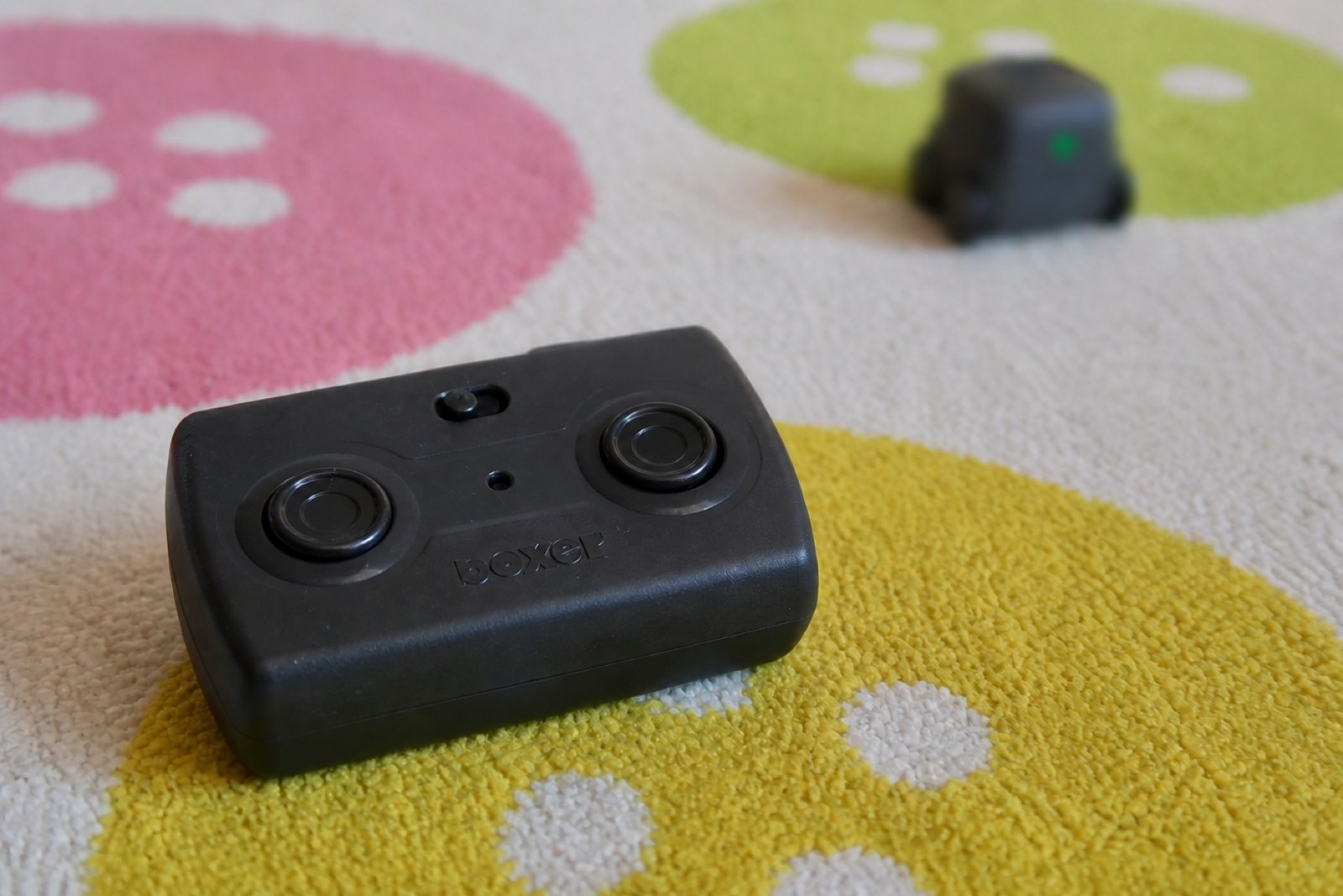 Close view of the charcoal grey Boxer remote control lying on a patterned rug, its two round black thumbsticks catching the light, with the powered on robot softly out of focus behind it.