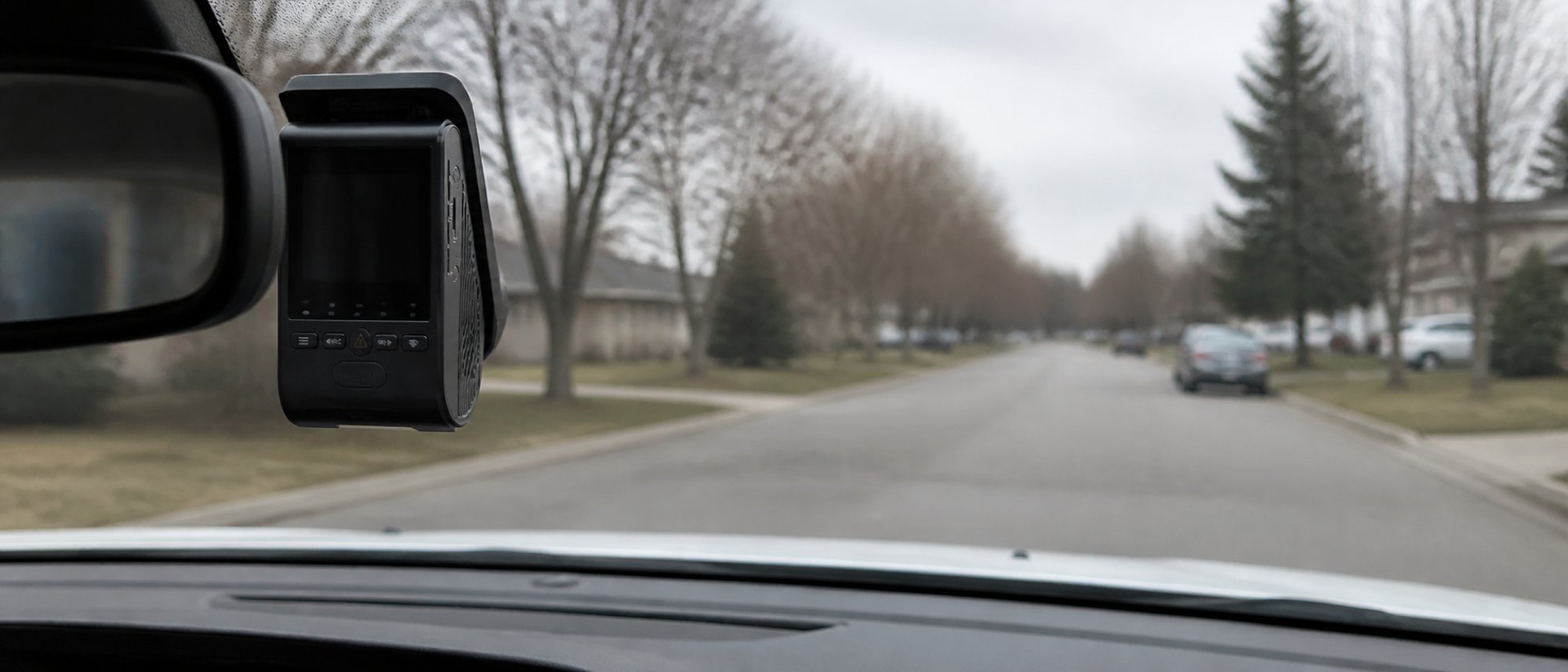 A small grey dash cam with a screen and a row of status lights mounted flush to a windshield just right of the rearview mirror, seen from the driver's seat on an overcast morning.