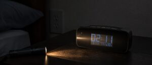 A flashlight beam raking sideways across a black digital alarm clock on a rental nightstand in a dim bedroom, with a wall outlet visible on the wall behind it.