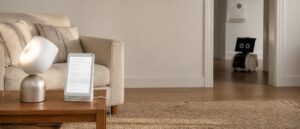 An ordinary living room with a faceless lamp shaped companion device and its docked tablet on a side table, and a knee height white robot on wheels parked in a doorway behind it.