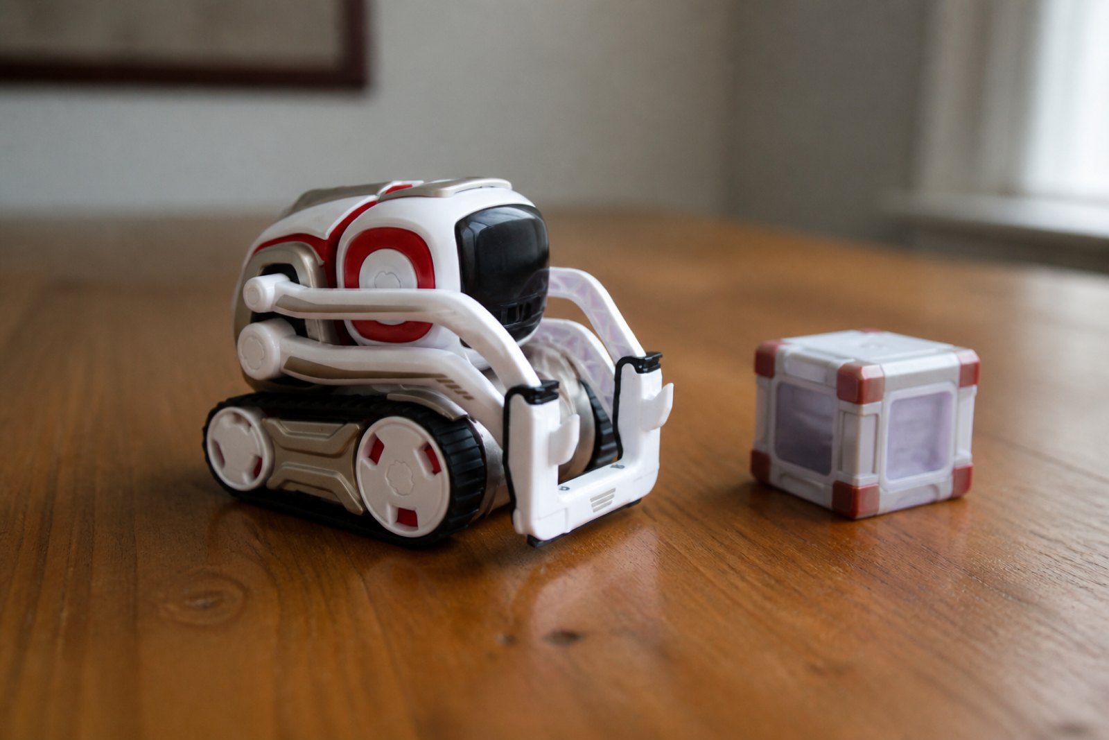 A white and red Cozmo robot on black rubber treads sitting on a wooden table, its glossy black face visor dark, one light up cube beside it.