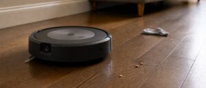 A charcoal grey disc shaped robot vacuum stopped on hardwood flooring, photographed at its own height, with the small square camera module visible low on its front bumper.
