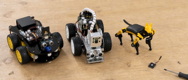 Overhead view of a workbench holding three hobby robot kits and one bare camera board: a black acrylic four wheel rover, a silver aluminum car with a pan and tilt camera bracket, a palm size yellow and black quadruped, and a postage stamp sized camera module with a stubby antenna.
