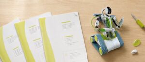 Overhead view of a workbench with three stapled A4 manuals in white and lime green fanned out beside a white and green tracked robot with a camera head on a gooseneck.